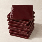 A stack of glossy square ceramic coasters in a deep burgundy red colour, arranged on a textured off-white surface.