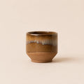A small ceramic matcha bowl with a brown and gold glaze, featuring a textured matte brown base and a glossy finish with a subtle white and blue-gray dripping pattern near the rim, set against a solid beige background.