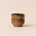 A small ceramic matcha bowl with a brown and gold glaze, featuring a textured matte brown base and a glossy finish with a subtle white and blue-gray dripping pattern near the rim, set against a solid beige background.