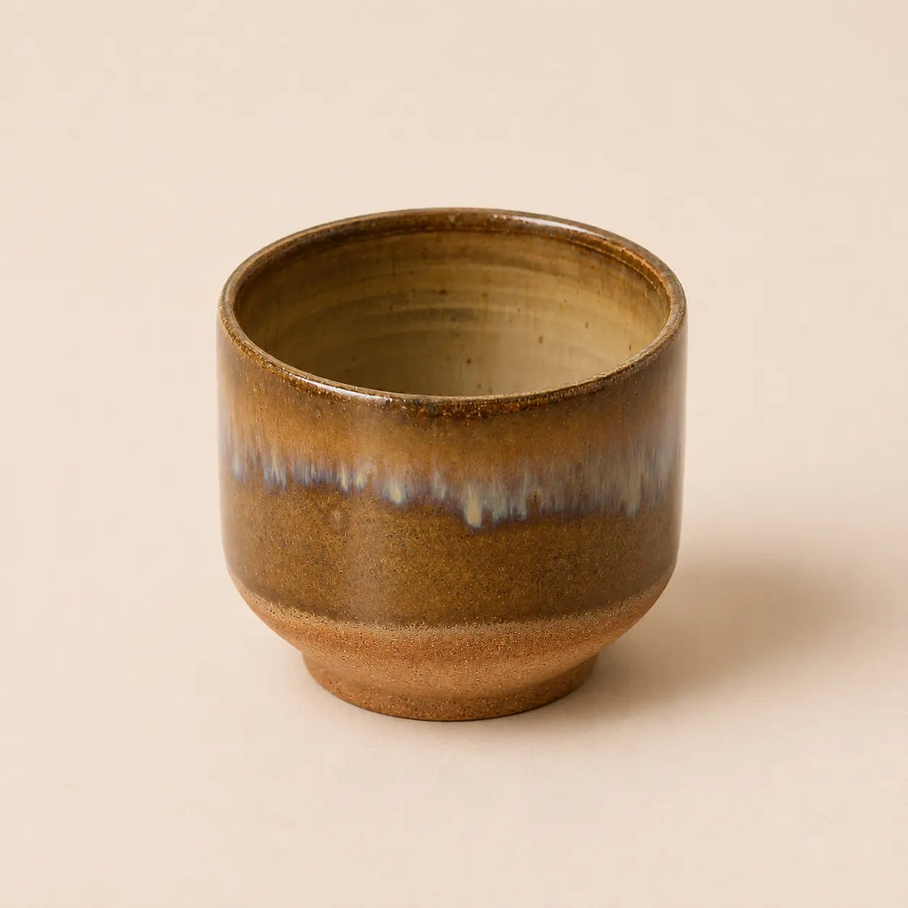 A small handmade ceramic matcha bowl with a brown and gold glaze and a blue-green dripping accent band, set against a solid beige background.