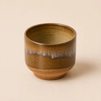 A small handmade ceramic matcha bowl with a brown and gold glaze and a blue-green dripping accent band, set against a solid beige background.