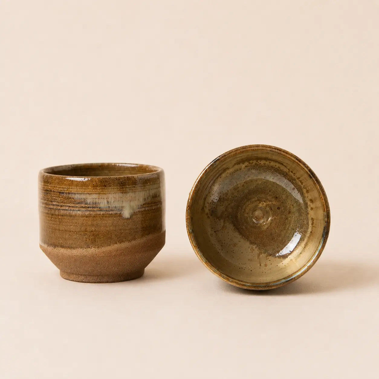 Two small handmade ceramic matcha bowl with a rustic brown and beige glaze, one standing upright and the other lying on its side, set against a solid light beige background.