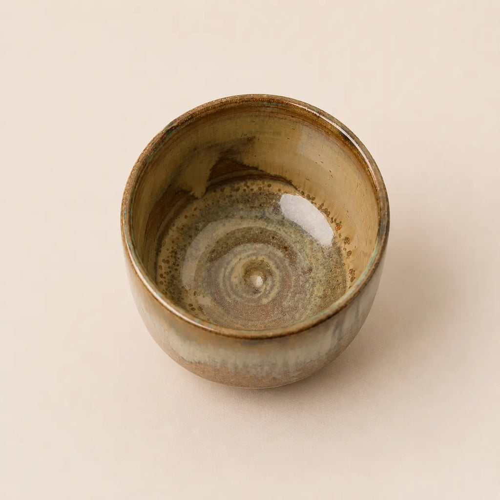 A handmade ceramic matcha bowl with a rustic spiral glaze pattern in earthy brown, beige, and muted green tones, photographed from a top-down perspective on a solid light beige background.