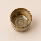 A handmade ceramic matcha bowl with a rustic spiral glaze pattern in earthy brown, beige, and muted green tones, photographed from a top-down perspective on a solid light beige background.