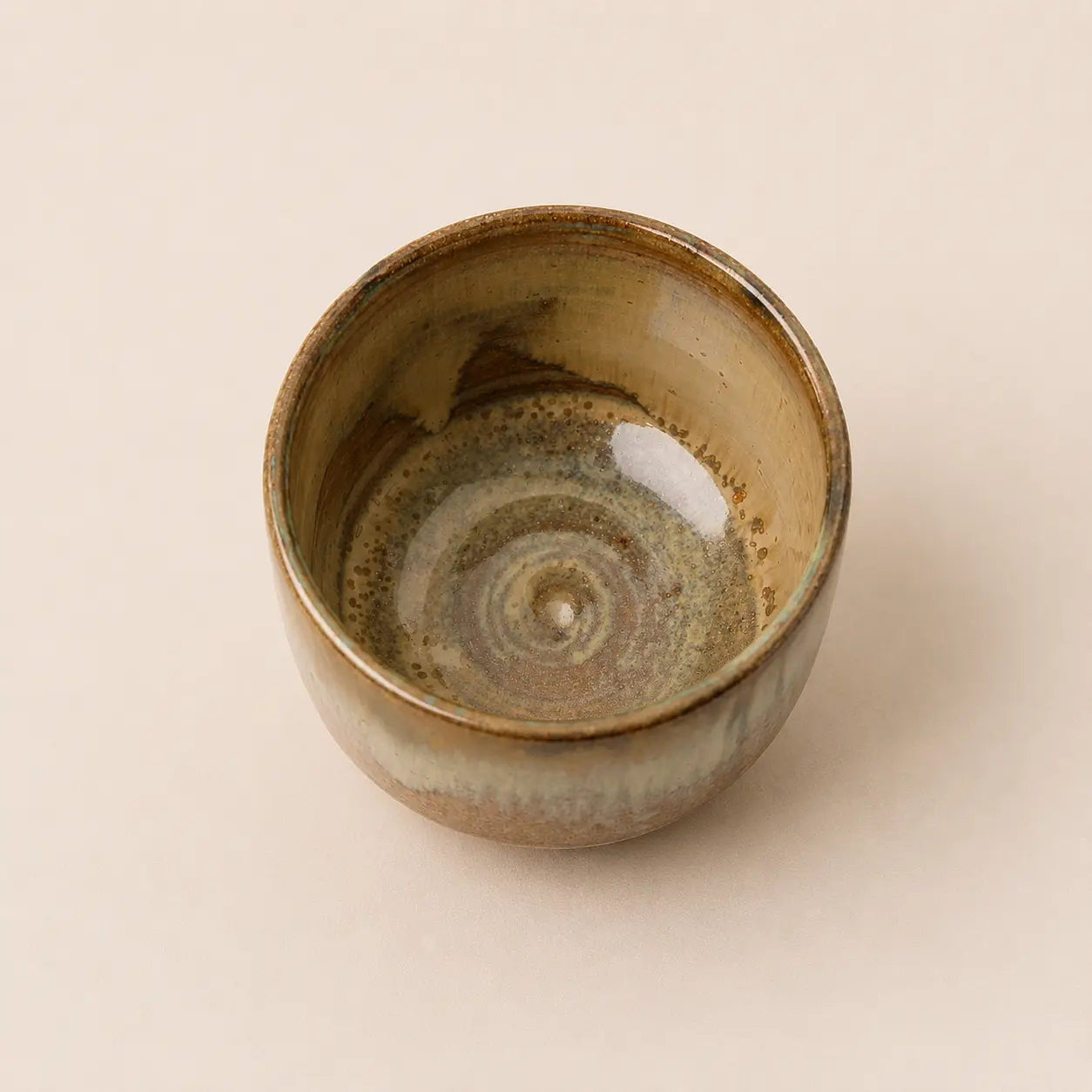 A handmade ceramic matcha bowl with a rustic spiral glaze pattern in earthy brown, beige, and muted green tones, photographed from a top-down perspective on a solid light beige background.