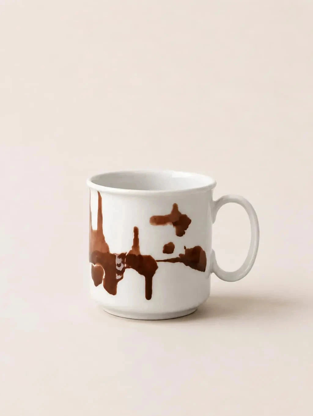 A white ceramic coffee mug with a brown dripping paint or chocolate splash design, set against a solid light beige background.
