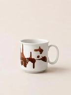 A white ceramic coffee mug with a brown dripping paint or chocolate splash design, set against a solid light beige background.