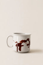 A white ceramic mug with a dark brown abstract paint-splatter design, set against a solid, warm off-white background.