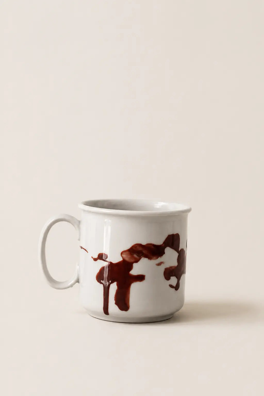 A white ceramic mug with a dark brown abstract paint-splatter design, set against a solid, warm off-white background.
