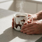 A white ceramic mug filled with a dark hot beverage, with steam rising, held by two hands on a light gray speckled countertop in a bright kitchen setting.