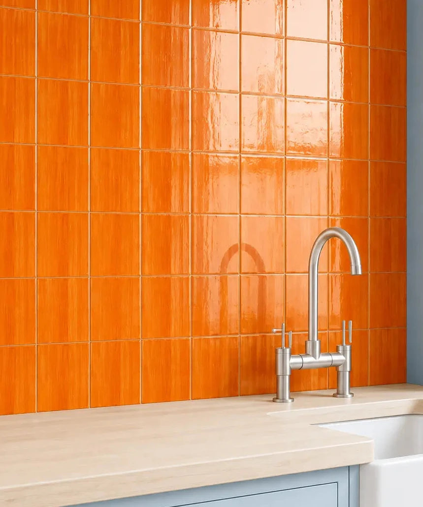 Bright orange glossy square wall tiles installed behind a light wood countertop and a chrome faucet, with pale blue cabinetry and a white farmhouse sink.
