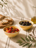 Two yellow ceramic snack bowls filled with food on a table: one with cherry tomatoes and the other with black olives, alongside sliced bread on a wooden board and a glass bottle of olive oil. The scene is styled with olive branches and soft natural shadows.