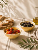 Two yellow ceramic snack bowls filled with food on a table: one with cherry tomatoes and the other with black olives, alongside sliced bread on a wooden board and a glass bottle of olive oil. The scene is styled with olive branches and soft natural shadows.