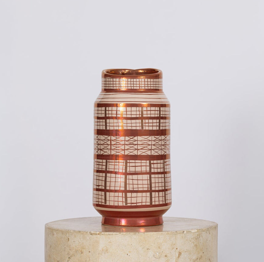 A small cylindrical vase with a copper-toned metallic finish and a white geometric grid pattern, displayed on a round beige pedestal against a light gray background.