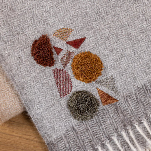 A close-up of a light gray woven throw blanket with cream fringe, featuring a terrazzo-style geometric pattern in rust, brown, beige, and gray, laid on a wooden surface.