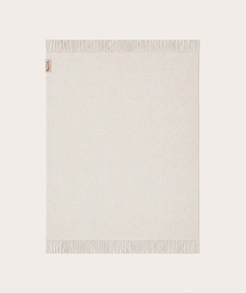 A light cream-coloured throw blanket with fringed edges, shown flat against a matching off-white background. A small red and white tag is visible on the top-left corner.