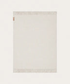 A light cream-coloured throw blanket with fringed edges, shown flat against a matching off-white background. A small red and white tag is visible on the top-left corner.