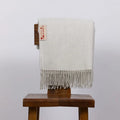 An off-white woven throw blanket with fringe, draped over a dark brown wooden stool. A small woven label on the blanket reads 'CARBON FIBRE'.