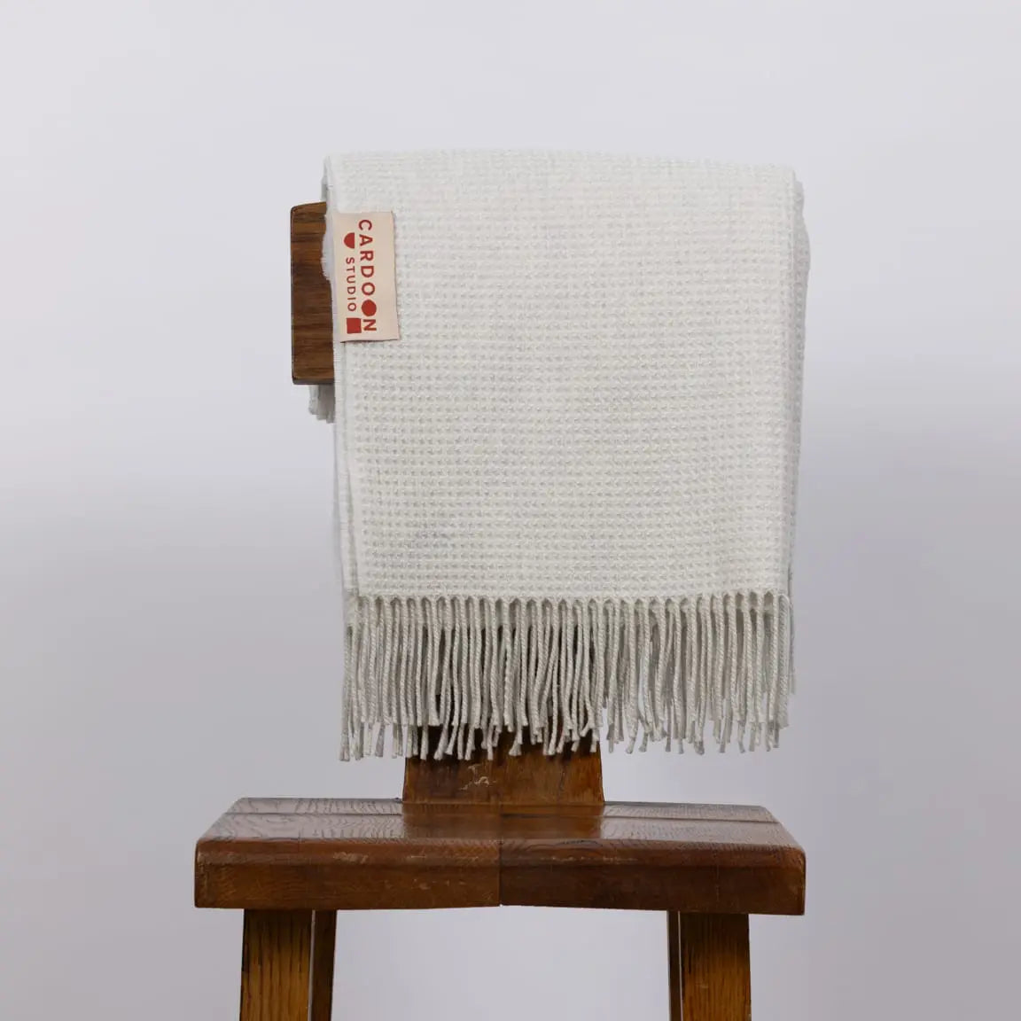 An off-white woven throw blanket with fringe, draped over a dark brown wooden stool. A small woven label on the blanket reads 'CARBON FIBRE'.