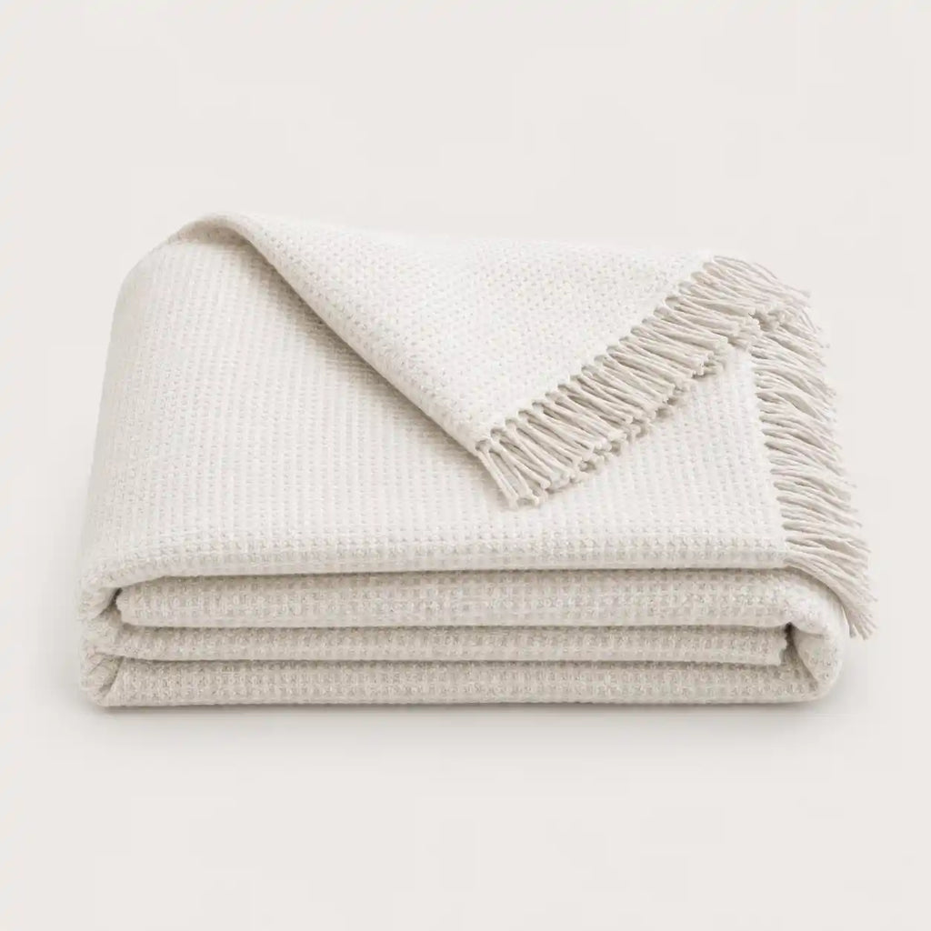 A folded off-white textured throw blanket with fringed edges, displayed on a clean, solid light background.