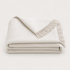 A folded off-white textured throw blanket with fringed edges, displayed on a clean, solid light background.