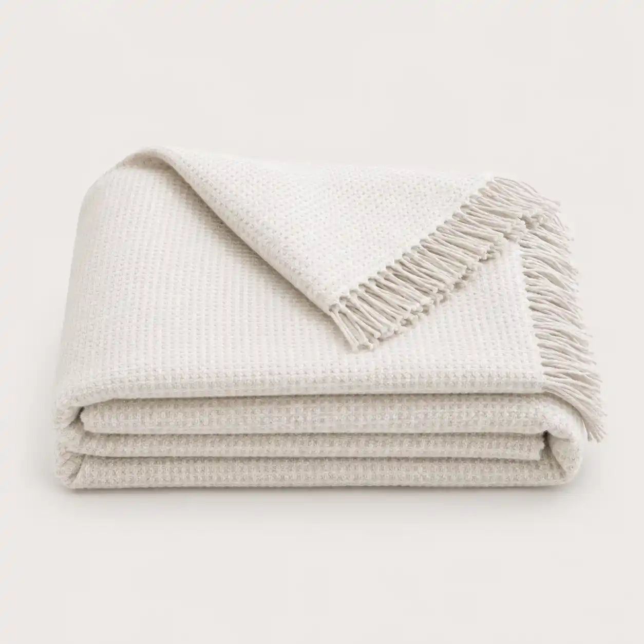 A folded off-white textured throw blanket with fringed edges, displayed on a clean, solid light background.