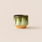 A small ceramic matcha bowl with a glossy green and white marbled glaze on the upper half, resting on a solid tan clay base against a warm beige background.