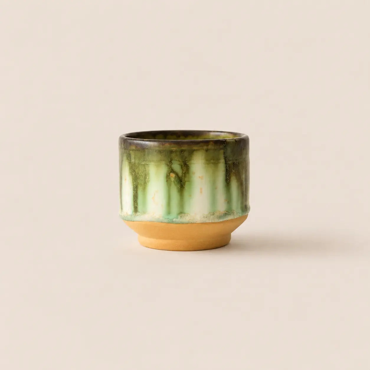 A small ceramic matcha bowl with a glossy green and white marbled glaze on the upper half, resting on a solid tan clay base against a warm beige background.