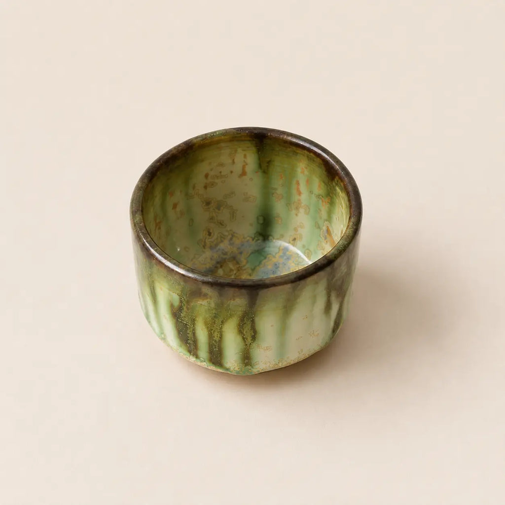 A small glazed ceramic matcha bowls with a vertical drip design in green and black against a solid beige background.