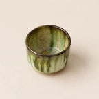A small glazed ceramic matcha bowls with a vertical drip design in green and black against a solid beige background.