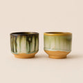 Two small glazed ceramic matcha bowls with a vertical drip design, one in green and black and the other in green and gold, set against a solid beige background.