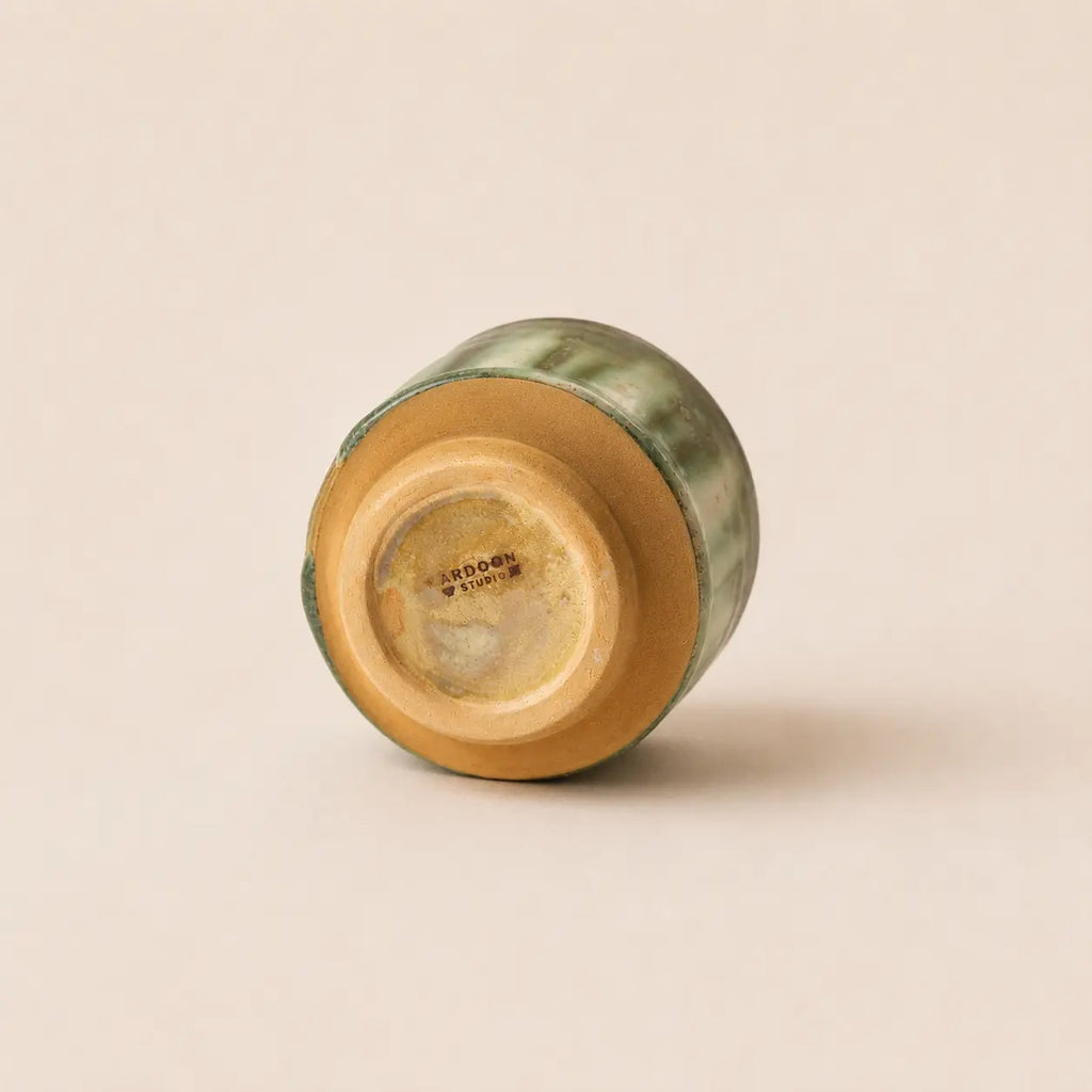 A small ceramic matcha bowl shown from its base, featuring a glossy green glaze and a natural tan clay foot, photographed on a solid light beige background.