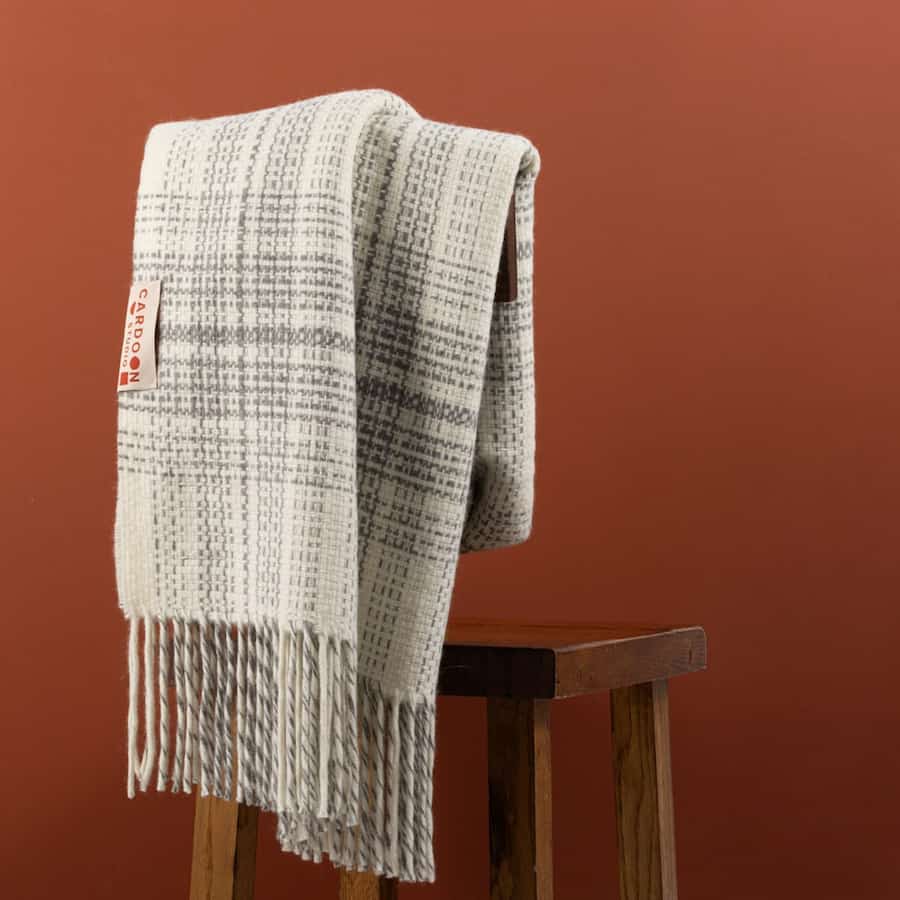 Soft woven alpaca blanket with subtle gray plaid texture draped over a wooden stool against a warm terracotta background.