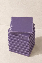 A stack of square ceramic lilac tiles in a muted lavender-purple glaze, with one coaster resting on top, set against a textured beige background.