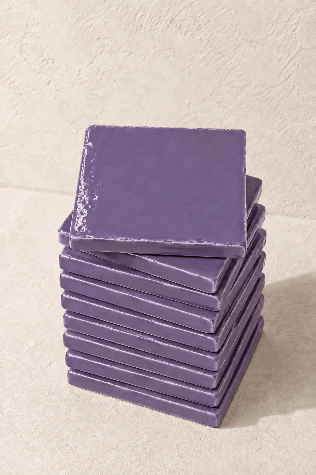 A stack of square ceramic lilac tiles in a muted lavender-purple glaze, with one coaster resting on top, set against a textured beige background.
