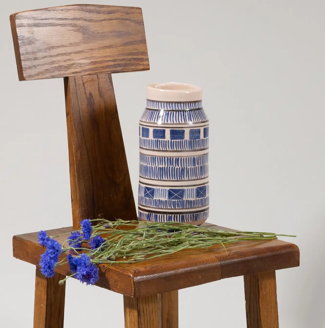 Blue patterned handcrafted ceramic vase displayed on a wooden chair alongside fresh blue flowers in a minimalist studio setting.