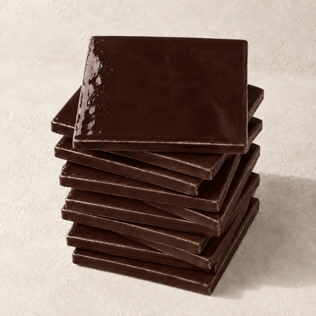A stack of glossy square ceramic tiles in a deep chocolate brown colour, arranged on a textured off-white surface.