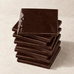 A stack of glossy square ceramic tiles in a deep chocolate brown colour, arranged on a textured off-white surface.