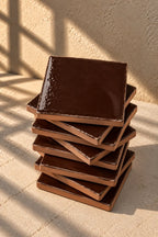 A stack of glossy square ceramic tiles in a deep chocolate brown colour, arranged on a textured off-white surface.