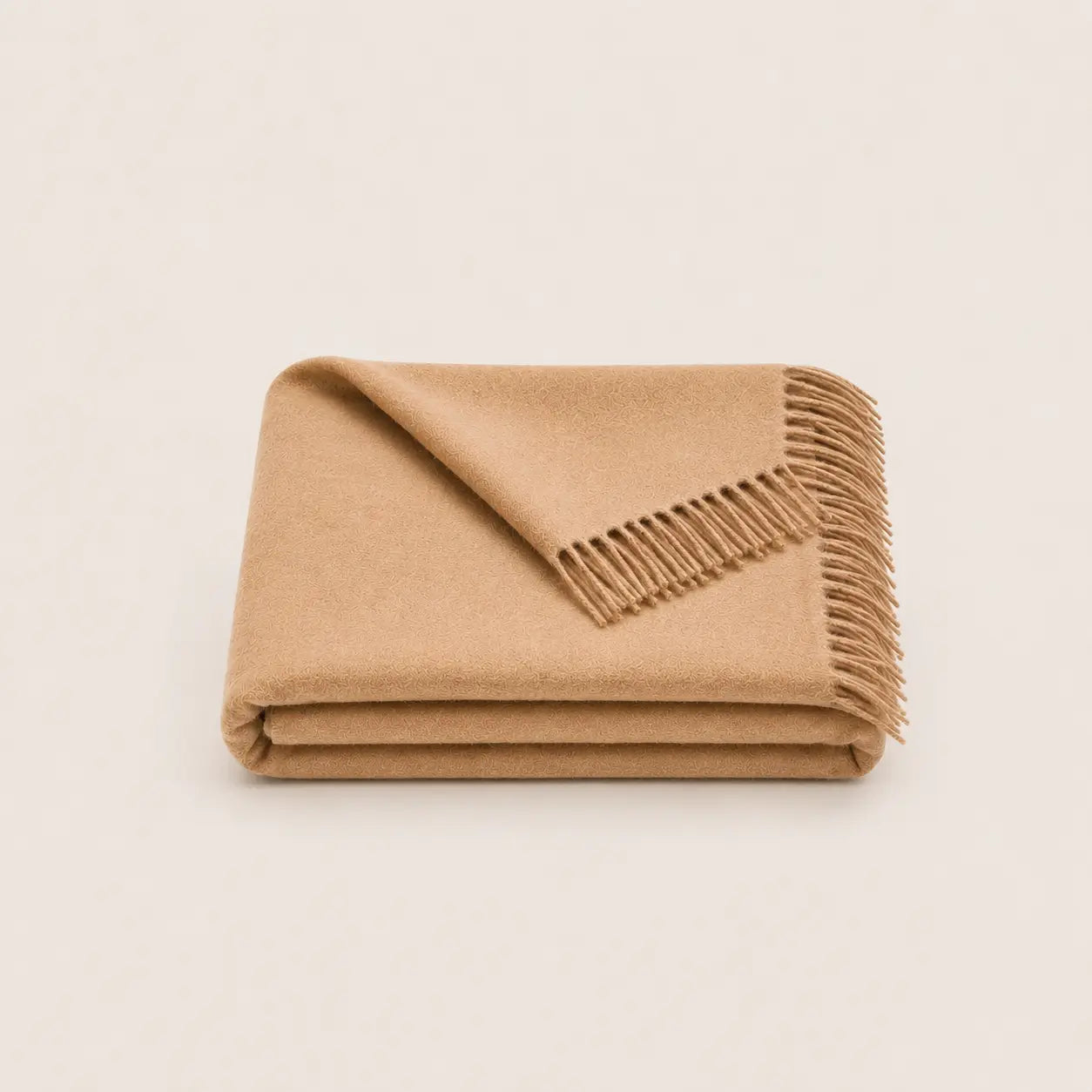 A folded camel colour alpaca throw blanket with fringed edges, presented on a clean, solid, light beige background.