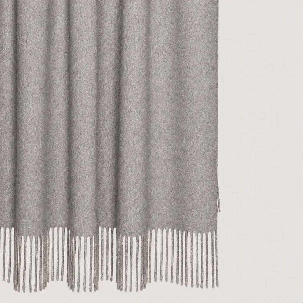 A close-up of a light gray textured throw blanket throw with fringe detailing along the bottom edge, shown against a plain off-white background.