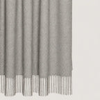A close-up of a light gray textured throw blanket throw with fringe detailing along the bottom edge, shown against a plain off-white background.
