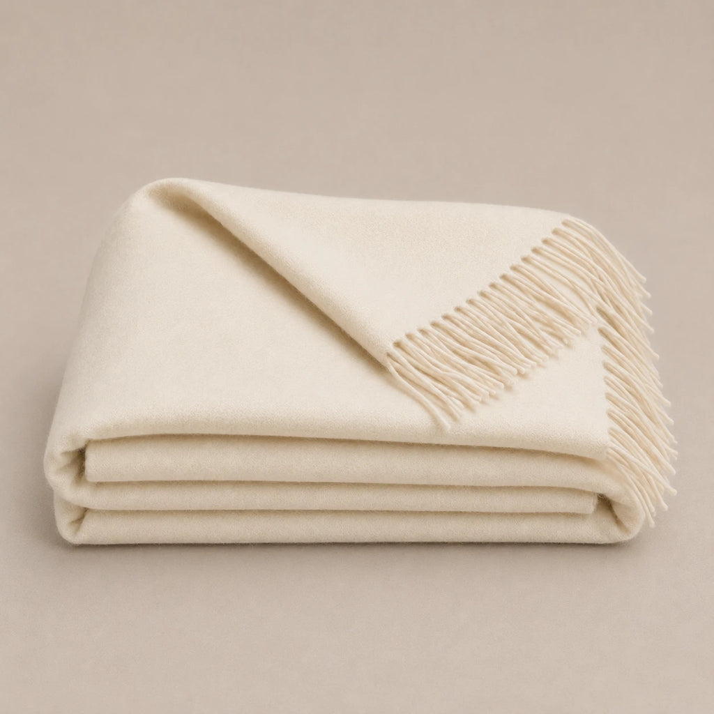A folded cream-colored alpaca throw blanket with fringed edges, displayed on a neutral beige background.