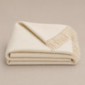 A folded cream-colored alpaca throw blanket with fringed edges, displayed on a neutral beige background.