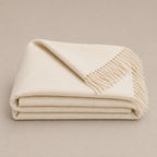 A folded cream-colored alpaca throw blanket with fringed edges, displayed on a neutral beige background.