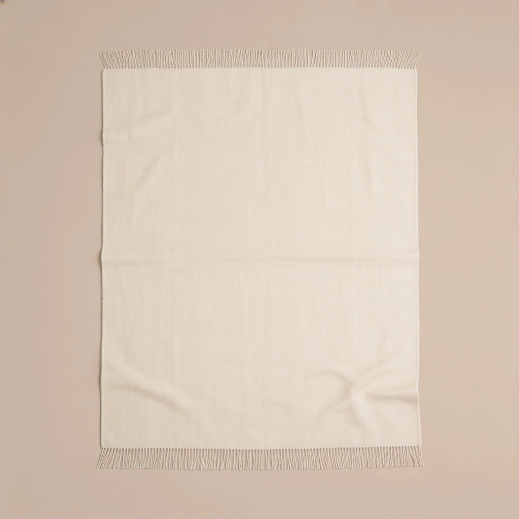 An off-white woven throw blanket with fringed edges, laid flat against a solid beige background.