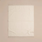 An off-white woven throw blanket with fringed edges, laid flat against a solid beige background.