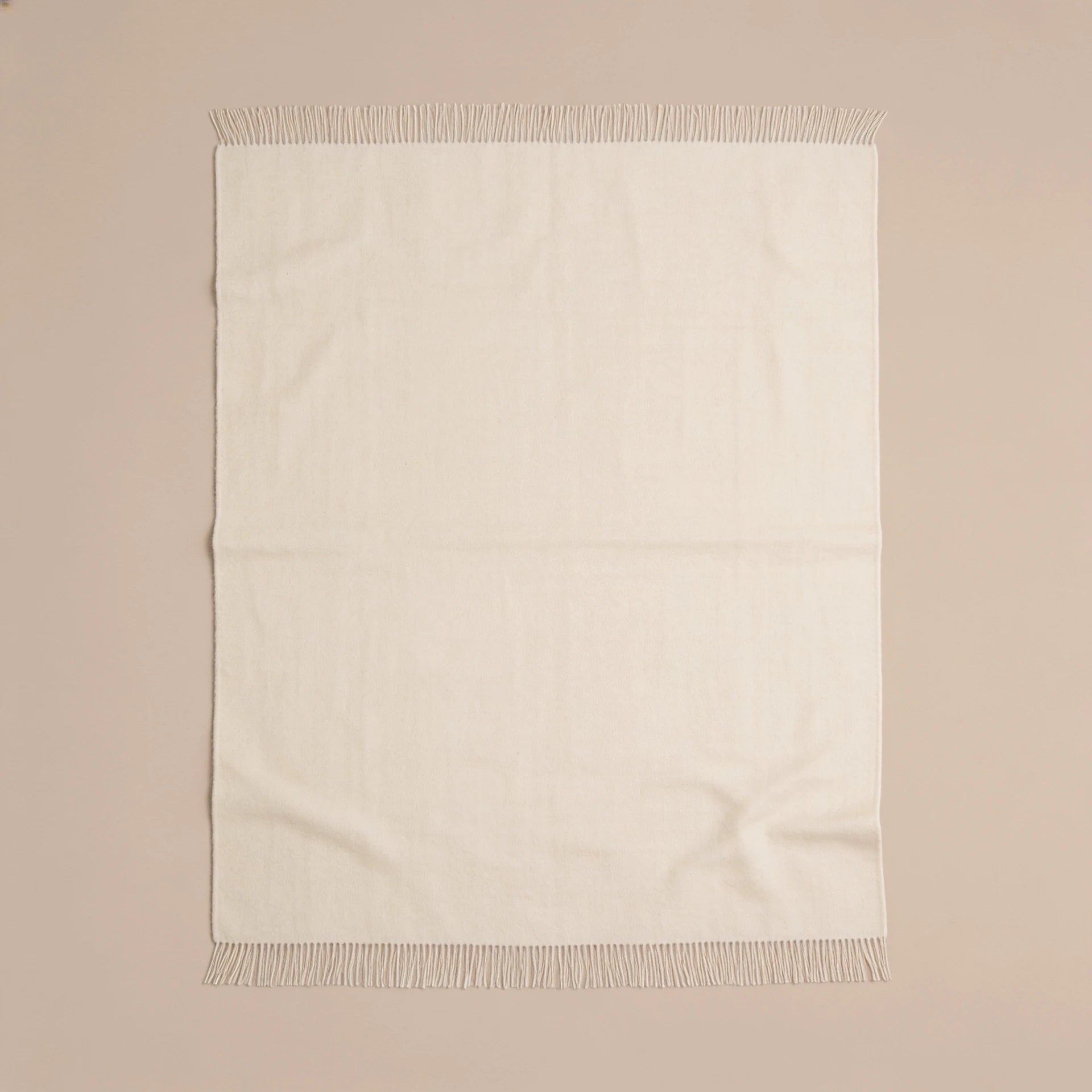 An off-white woven throw blanket with fringed edges, laid flat against a solid beige background.