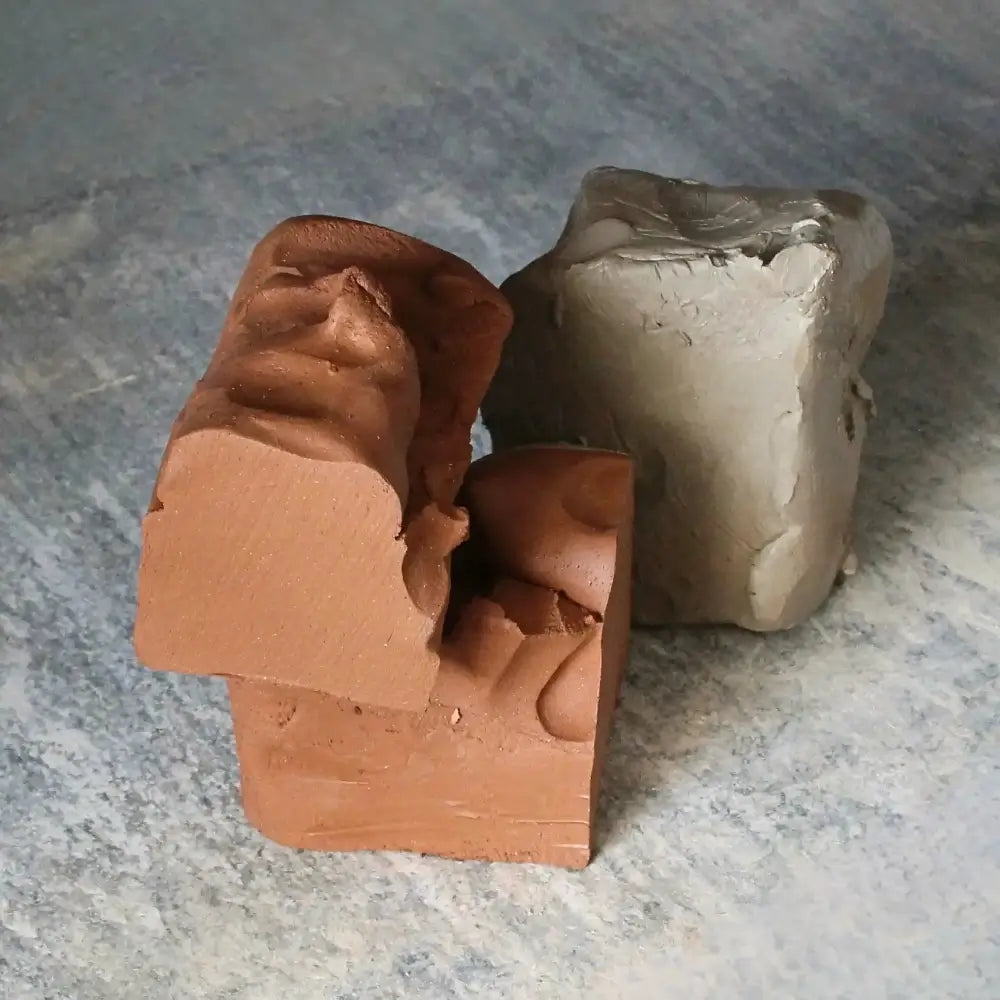 Two blocks of clay, one reddish-brown and one gray, placed on a textured surface.