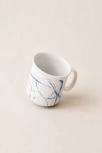 A white ceramic mug with a blue abstract line pattern, photographed on a solid, warm off-white background.